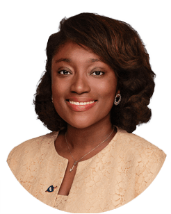 A woman with shoulder-length curly hair, wearing a beige lace blazer, a necklace, and earrings, smiling at the camera against a plain background.