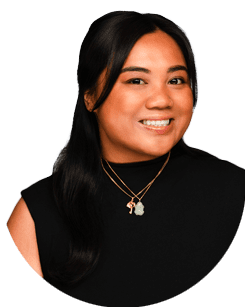 Woman with long dark hair wearing a sleeveless black top and layered gold necklaces, smiling at the camera against a plain light background.