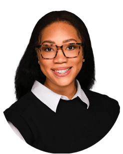 A woman with long dark hair, wearing tortoiseshell glasses, a white collared shirt, and a black top, smiles at the camera against a plain background.