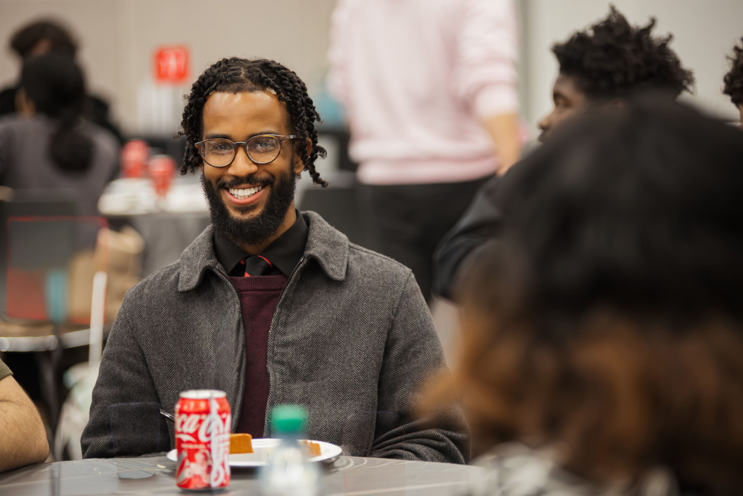 A man with glasses and a beard smiles while sitting at a table with a plate of food and a can of Coca-Cola in front of him. Other people are seated around him.