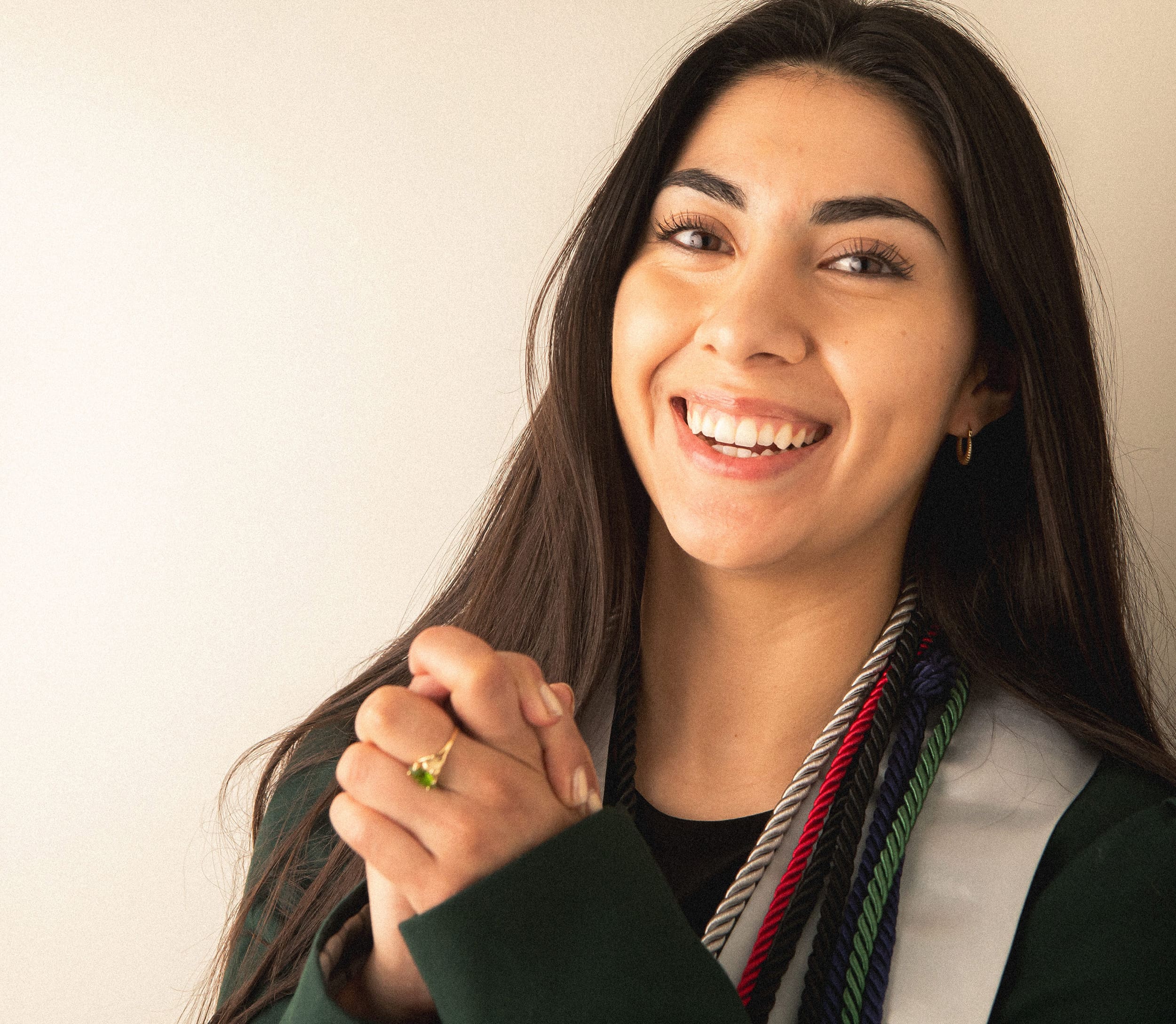 Young person with long dark hair smiling and clasping her hands, wearing a green jacket and multi-colored cords draped around her neck, standing against a plain background.