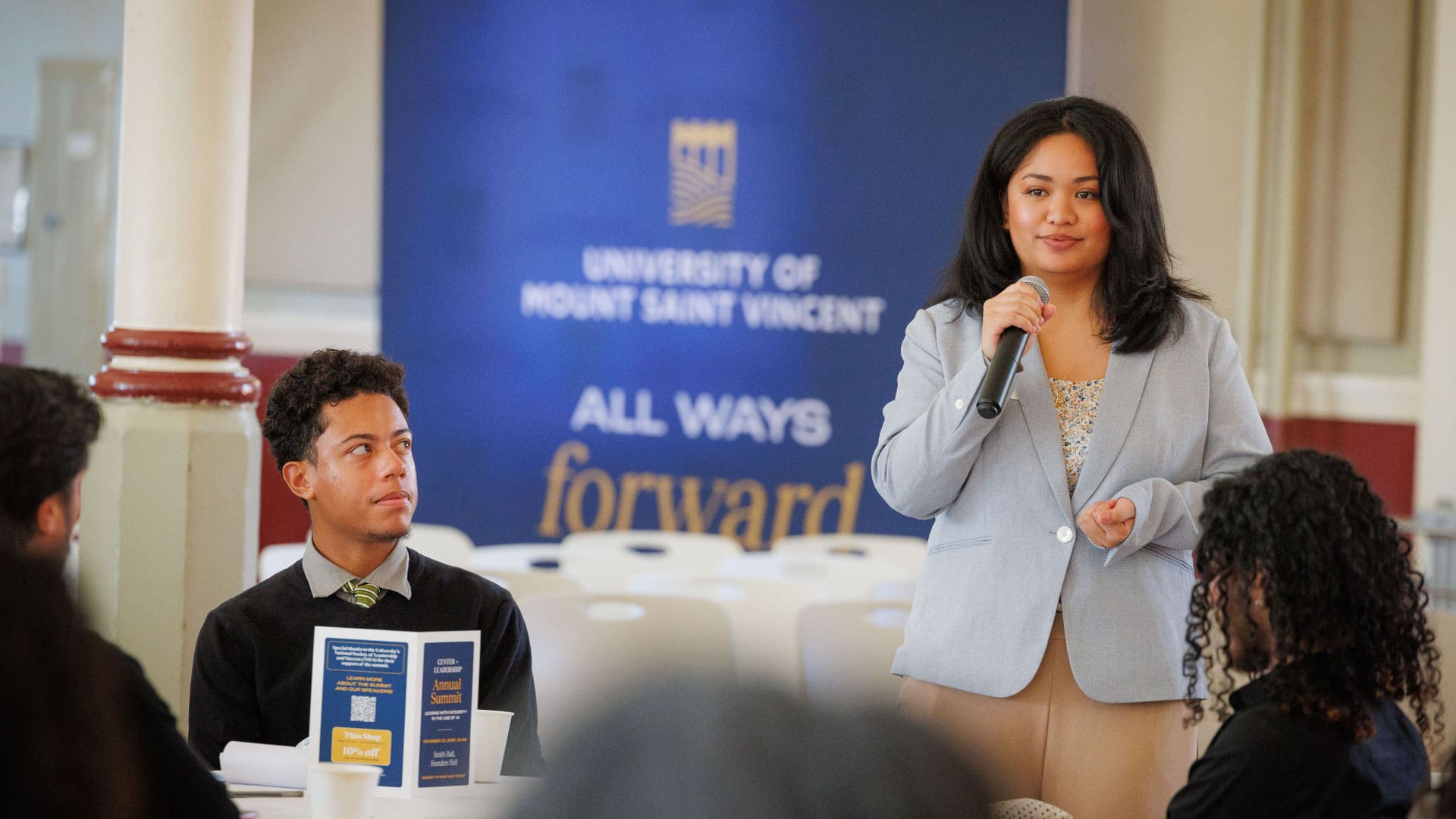 A woman speaks into a microphone at a table with others while a University of Mount Saint Vincent banner is displayed in the background.