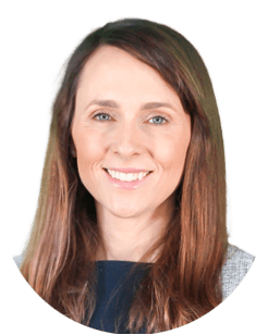 A woman with long brown hair and light skin is smiling at the camera, wearing a dark top and a gray jacket, against a plain light background.