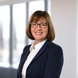 A woman wearing glasses and a navy blazer over a white shirt smiles at the camera in a brightly lit office setting.
