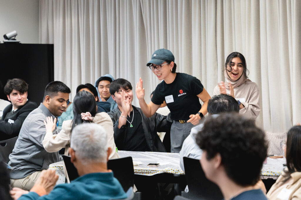 A group of people sit and stand around a table indoors; some are smiling and giving high fives, while others watch and chat.