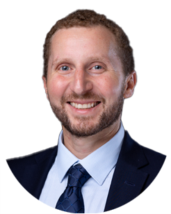 A smiling man with short curly hair and a beard, wearing a dark suit, light blue shirt, and blue tie, in front of a plain white background.