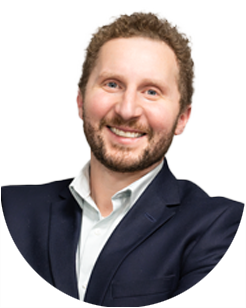 Smiling man with curly hair and beard wearing a dark suit jacket and light shirt, posed against a plain background.