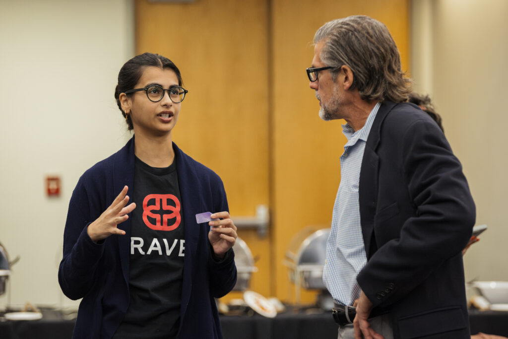 Two people have a conversation indoors; one gestures while wearing glasses and a shirt with "RAVE" on it, the other listens attentively. Buffet trays are visible in the background.