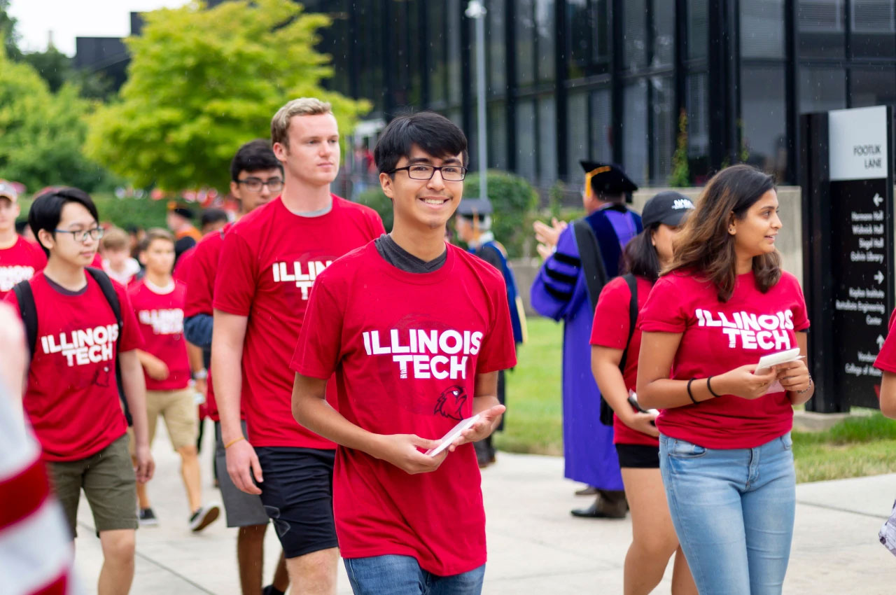 A group of students wearing red Illinois Tech t-shirts walk together outdoors on a campus pathway, with trees and buildings in the background.