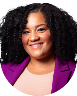 Woman with curly black hair wearing a purple blazer and light pink top, smiling at the camera against a light background.