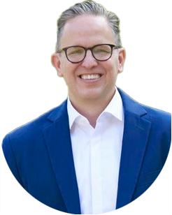 A person wearing glasses, a white shirt, and a blue blazer smiles at the camera against a plain background.