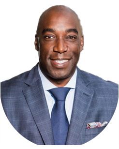 A man in a blue checked suit, light blue shirt, and navy tie, smiling at the camera against a plain light background.