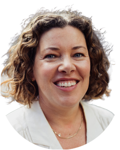 Smiling woman with curly brown hair wearing a white top and a gold necklace, facing the camera against a plain background.