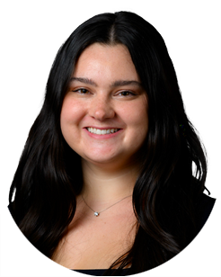 A woman with long dark hair, wearing a black top and a necklace, smiling at the camera against a plain white background.
