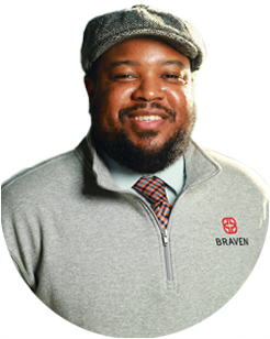A man wearing a gray flat cap, gray zip-up sweater with a Braven logo, dress shirt, and plaid tie, smiling at the camera.