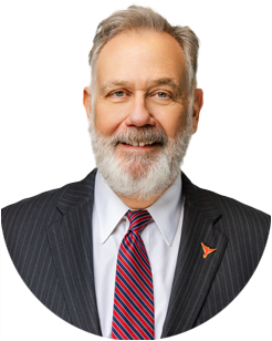 A man with gray hair and a beard wearing a dark pinstripe suit, white shirt, and red striped tie, posing and smiling at the camera.