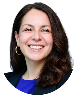 Woman with long brown hair, wearing a blue top, black blazer, and gold hoop earrings, smiling and looking slightly to her left.