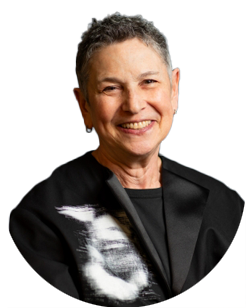 Smiling older person with short gray hair, wearing a black jacket over a dark shirt, shown from the chest up against a plain light background.