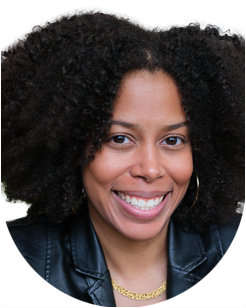 Woman with curly hair smiles at the camera, wearing a black leather jacket, gold hoop earrings, and a gold necklace, against a light background.