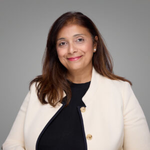 A woman with long brown hair wearing a cream blazer over a black top smiles at the camera against a plain gray background.