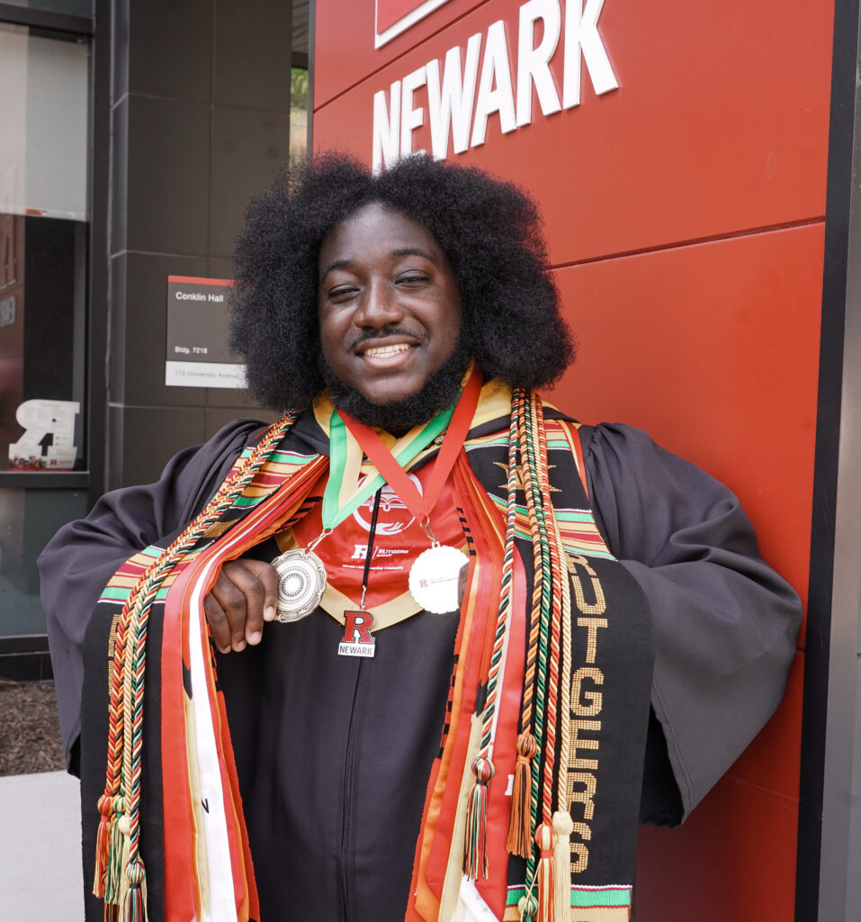 A graduate in a gown and stole stands in front of a Rutgers Newark sign, wearing multiple medals and cords, and smiling at the camera.