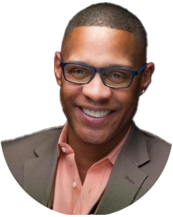 Smiling man wearing glasses, a brown blazer, and a light pink shirt, pictured from the shoulders up against a light background.