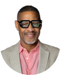 Smiling man with short hair, beard, and mustache wears black glasses, a light beige blazer, and a pink collared shirt, pictured against a plain light background.