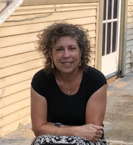 A woman with curly hair, wearing a black shirt and patterned skirt, sits outside near a tan siding wall and a glass-paneled door, smiling at the camera.