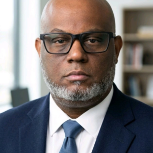A bald man with glasses, a greying beard, and a navy suit with a tie poses in an office setting with shelves and windows in the background.