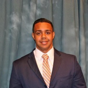 A man in a dark suit, white shirt, and striped tie stands in front of a blue curtain, smiling at the camera.