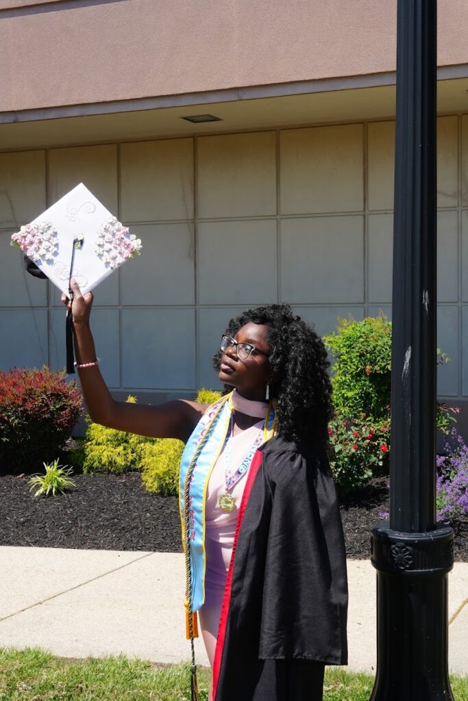 A graduate in a gown and honor cords holds up a decorated graduation cap outdoors near a building and a lamp post.