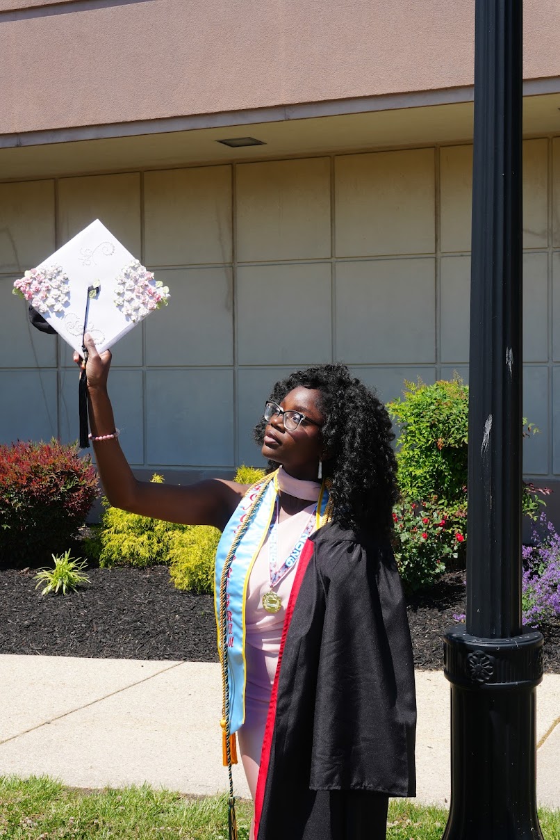A graduate in a gown and honor cords holds up a decorated graduation cap outdoors near a building and a lamp post.