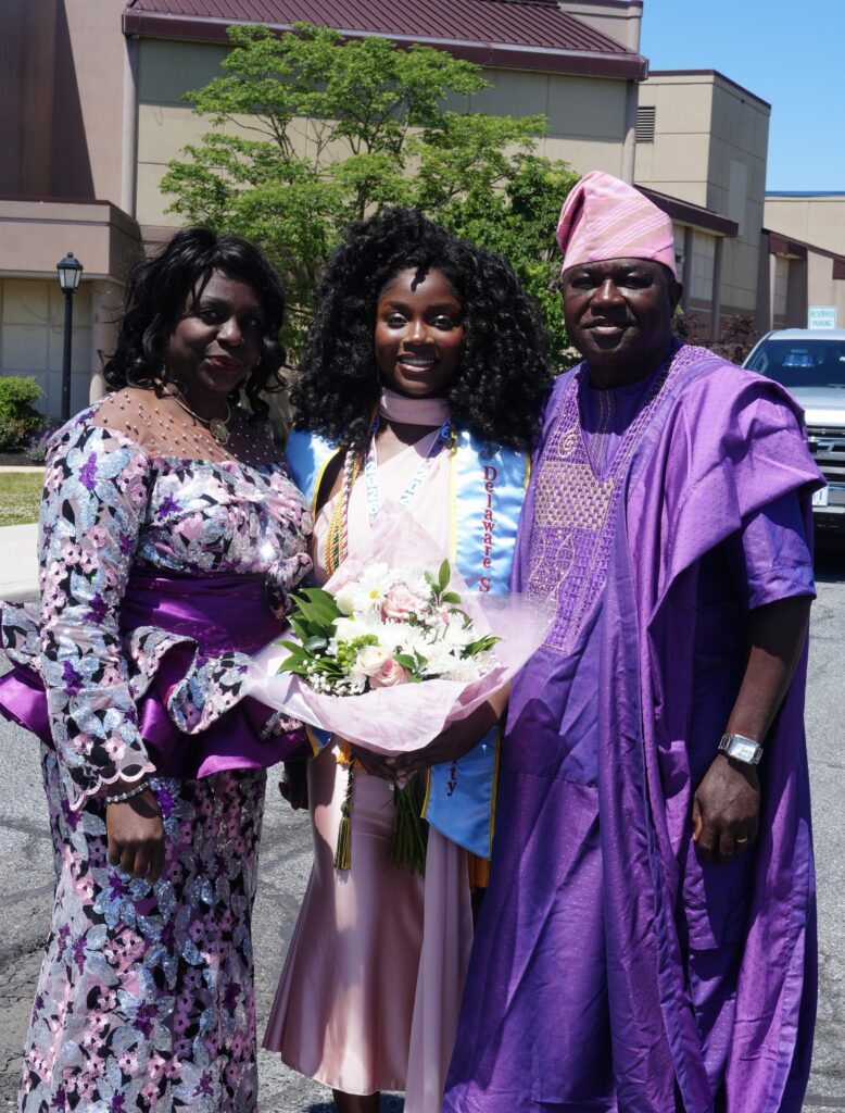 Three people stand outdoors; one young woman in graduation attire holds flowers, flanked by two adults in colorful formal clothing.