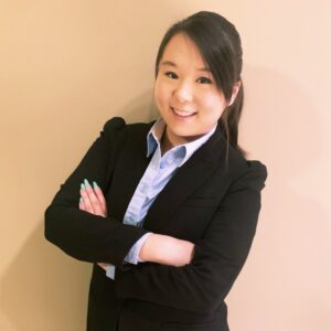 Woman in a black blazer and blue shirt standing against a beige wall with arms crossed, smiling at the camera.