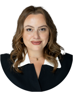 Woman with wavy brown hair wearing a black jacket and white-collared shirt, smiling at the camera against a plain light background.
