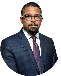 A man with a goatee and glasses wearing a navy suit, white shirt, and maroon tie.